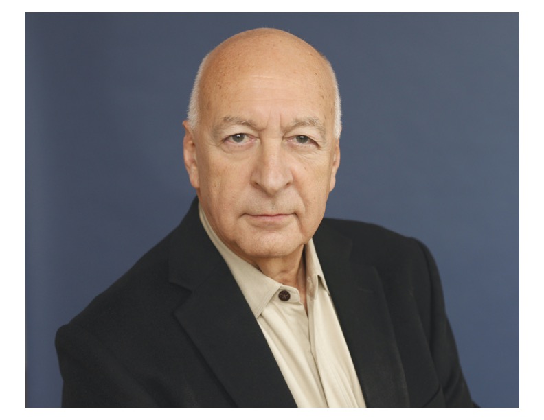 Portrait of a person with white hair wearing a black jacket over white button down in front of a blue backdrop