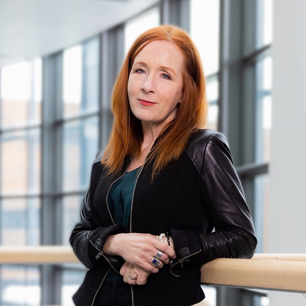 Portrait of a person with mid-length orange hair wearing a black jacket over a green top leaning against a brown railing in a glass building