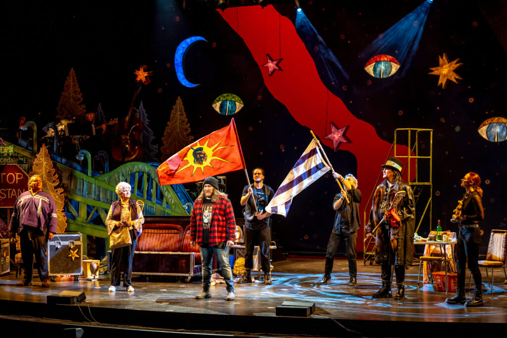 Still of a theatre production of Beautiful Scars with seven cast members on stage, two of them holding up large flags in front of a colourful set.