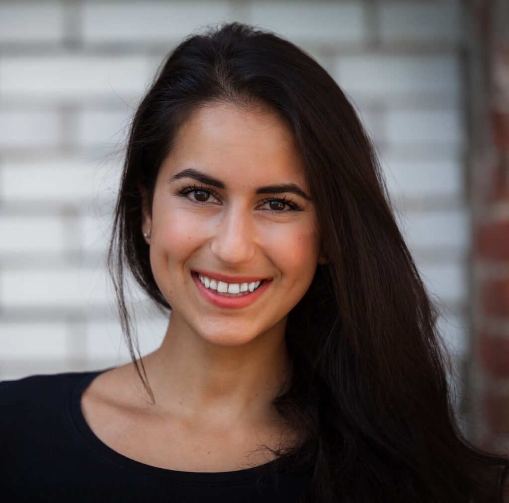 Woman with long dark hair and black shirt smiling in front of white brick wall