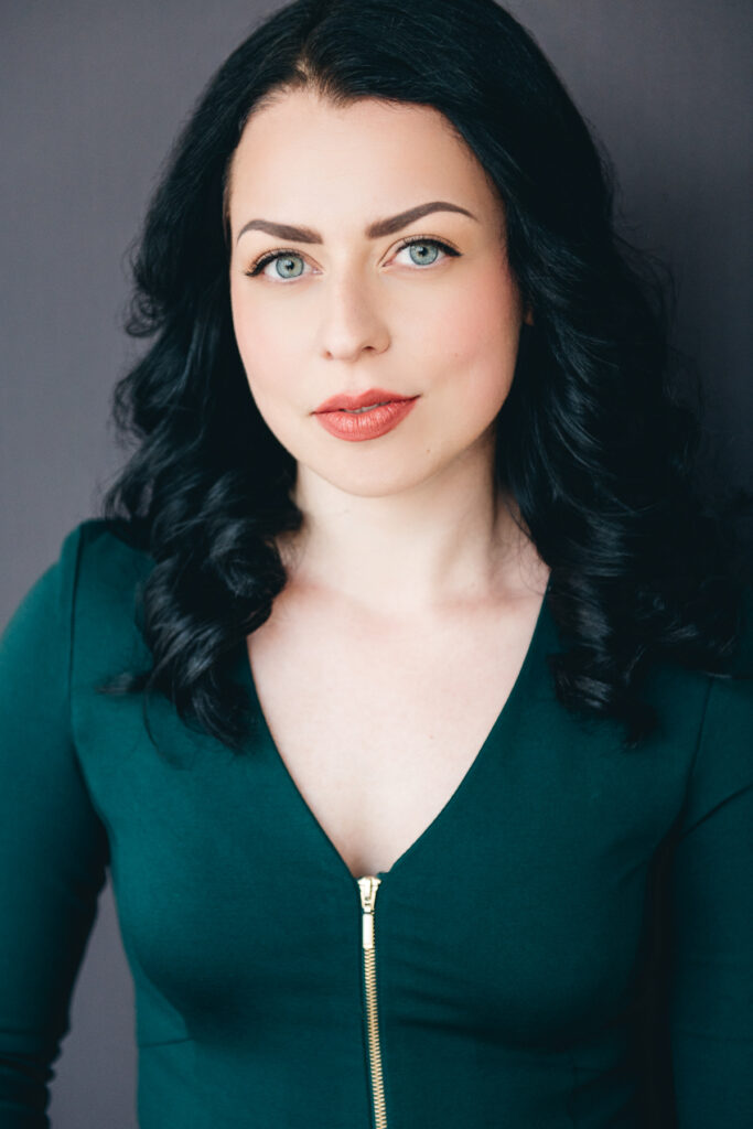 Portrait of person with long curly blue-black hair and dark green v-neck top in front of a gray background