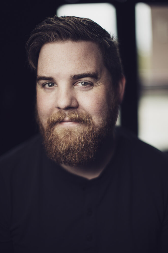 Portrait of a person with dark bond mustache and beard wearing a dark top, posing in front of a window. Photo by David Howells