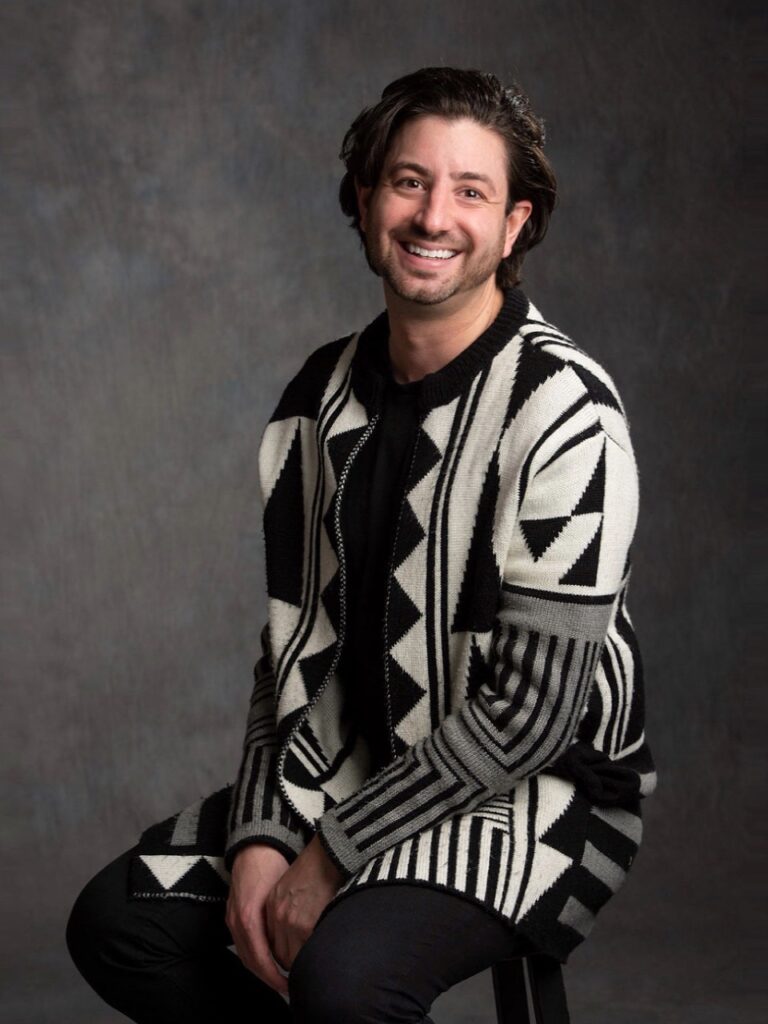 Half body studio portrait of person with curled dark hair wearing a long black and white patterned top.