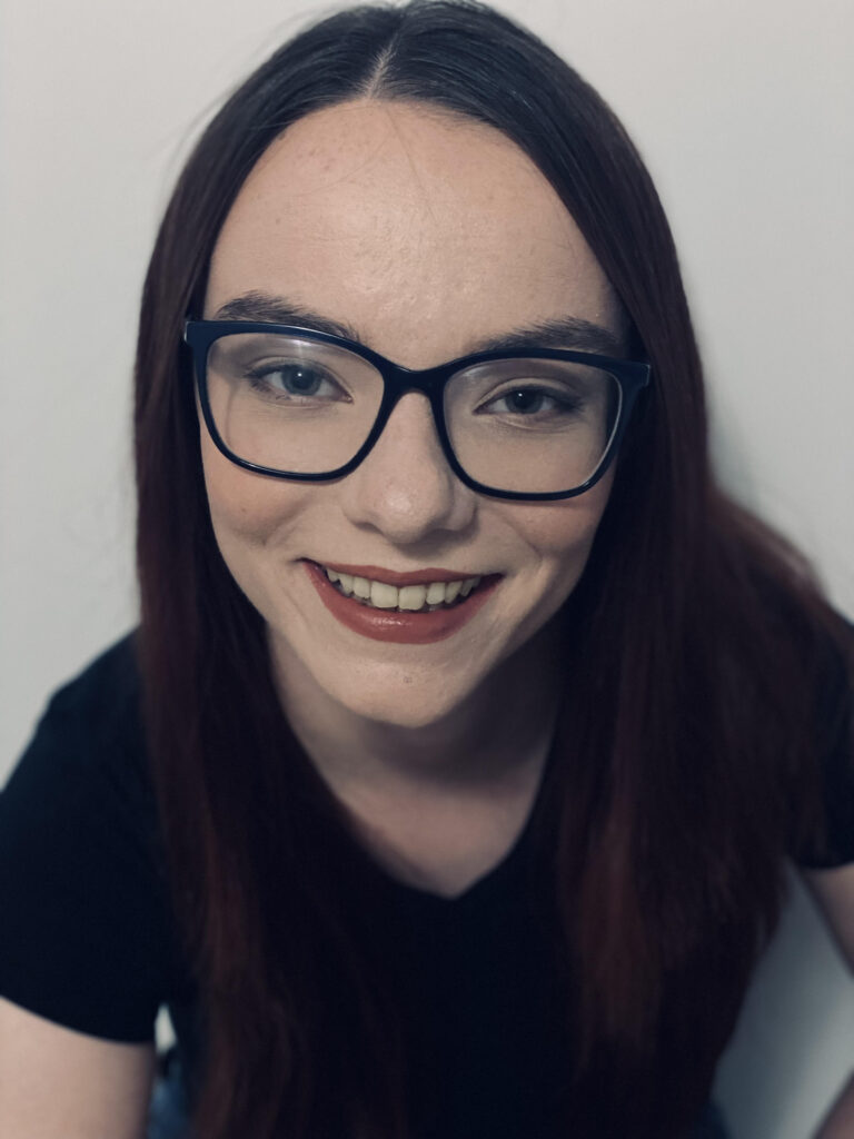 Portrait of person with straight long dark hair, black shirt, large dark-framed glasses, and wearing red lipstick