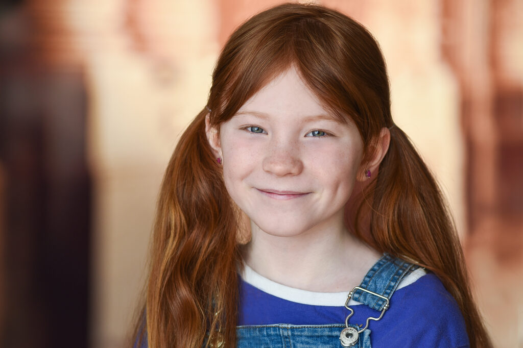 Portrait of a young child with long red hair in ponytails wearing denim overalls and blue and white top.