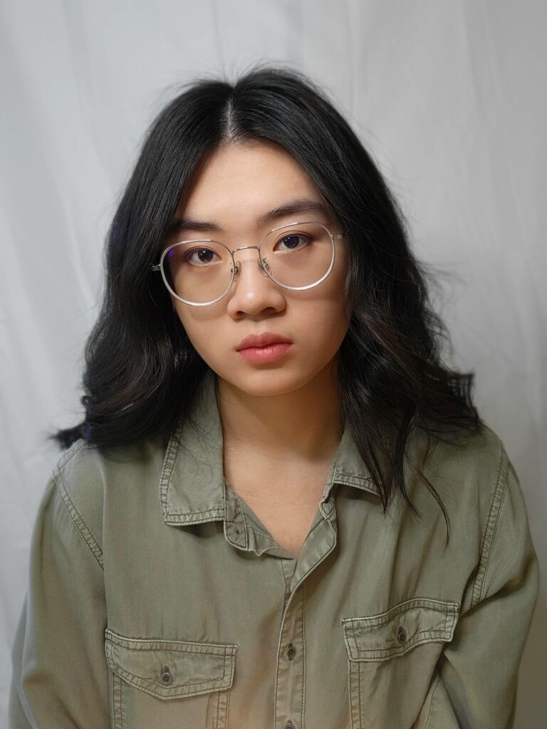 Woman with dark hair and glasses and green button up in front of white fabric wall