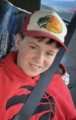 Picture of child with short brown hair wearing a red and white baseball cap, red hoodie, and sitting in a car