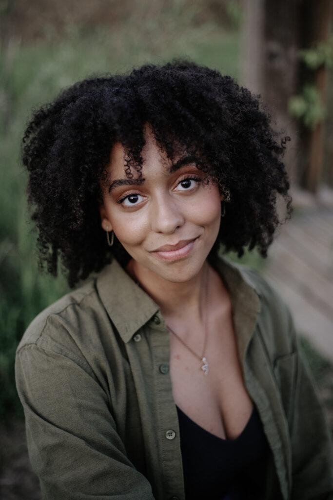 Portrait of a person with long dark afro, black top, and open green button down shirt