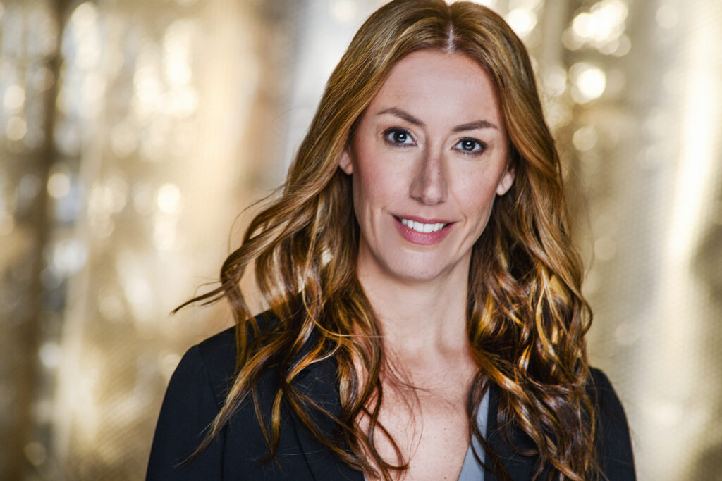 Portrait of a person with long wavy dirty blonde hair wearing a gray top and black coat in front of a glittery gold background