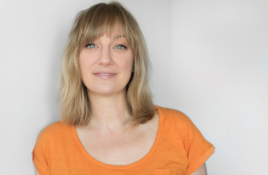 Portrait of a person with mid-length blonde hair and blunt bangs, wearing an orange shirt in front of a white background