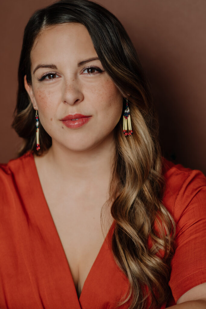 Portrait of a person with long brown hair, orange v-neck top, dangling earrings, and orange background