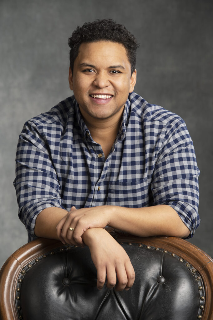 Studio portrait of person with spiky dark hair, black and white check shirt, with arms leaning against the back of a dark leather chair