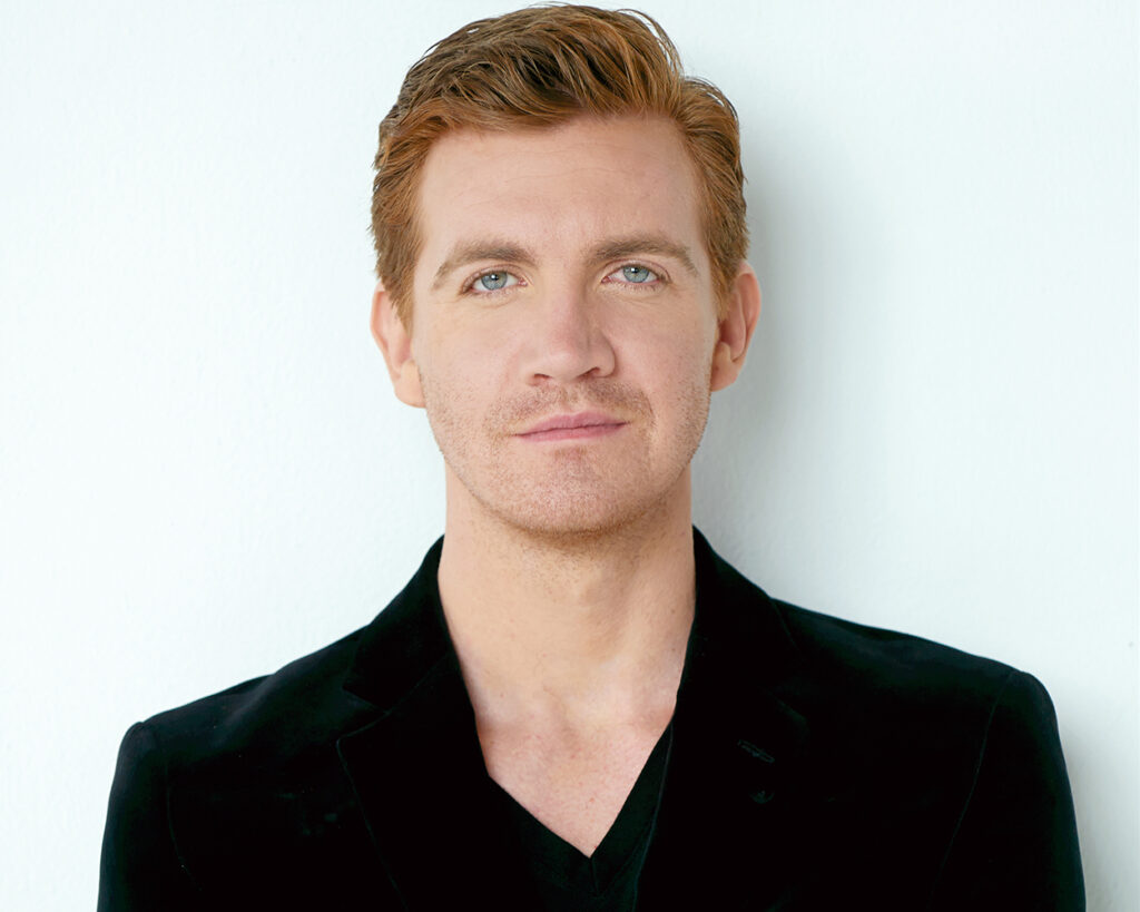 Portrait of a person with quiffed orange hair wearing a black top in front of a white background