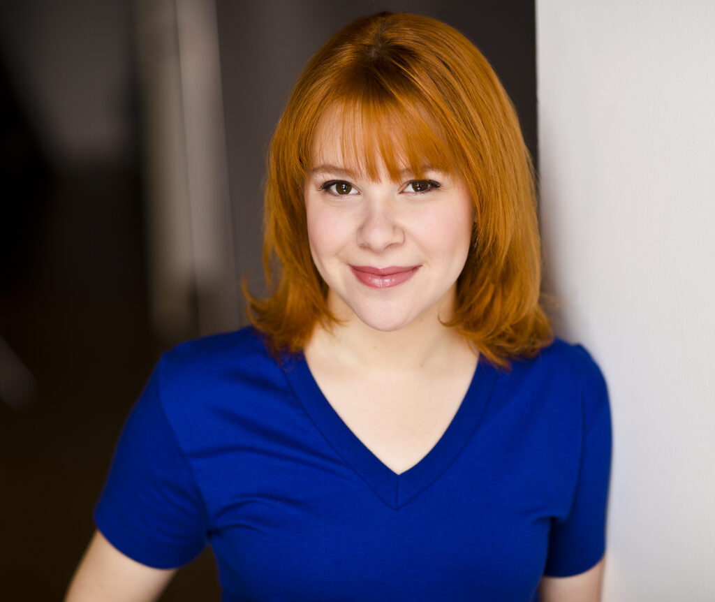 Portrait of a young person with shoulder length orange hair and bangs wearing a blue short-sleeve top leaning against a white wall