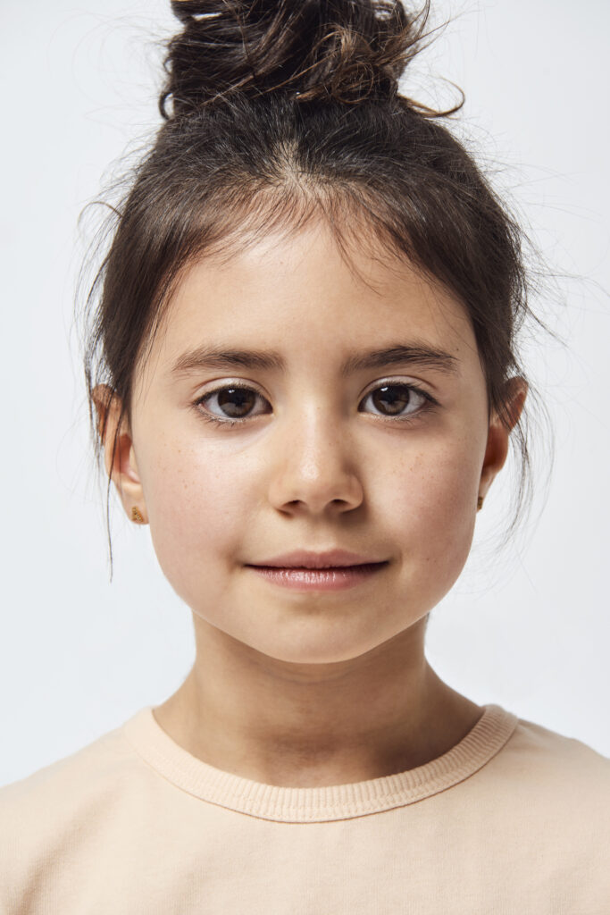 Studio portrait of little girl with dark hair in a bun at the top pf her head wearing a yellow shirt.