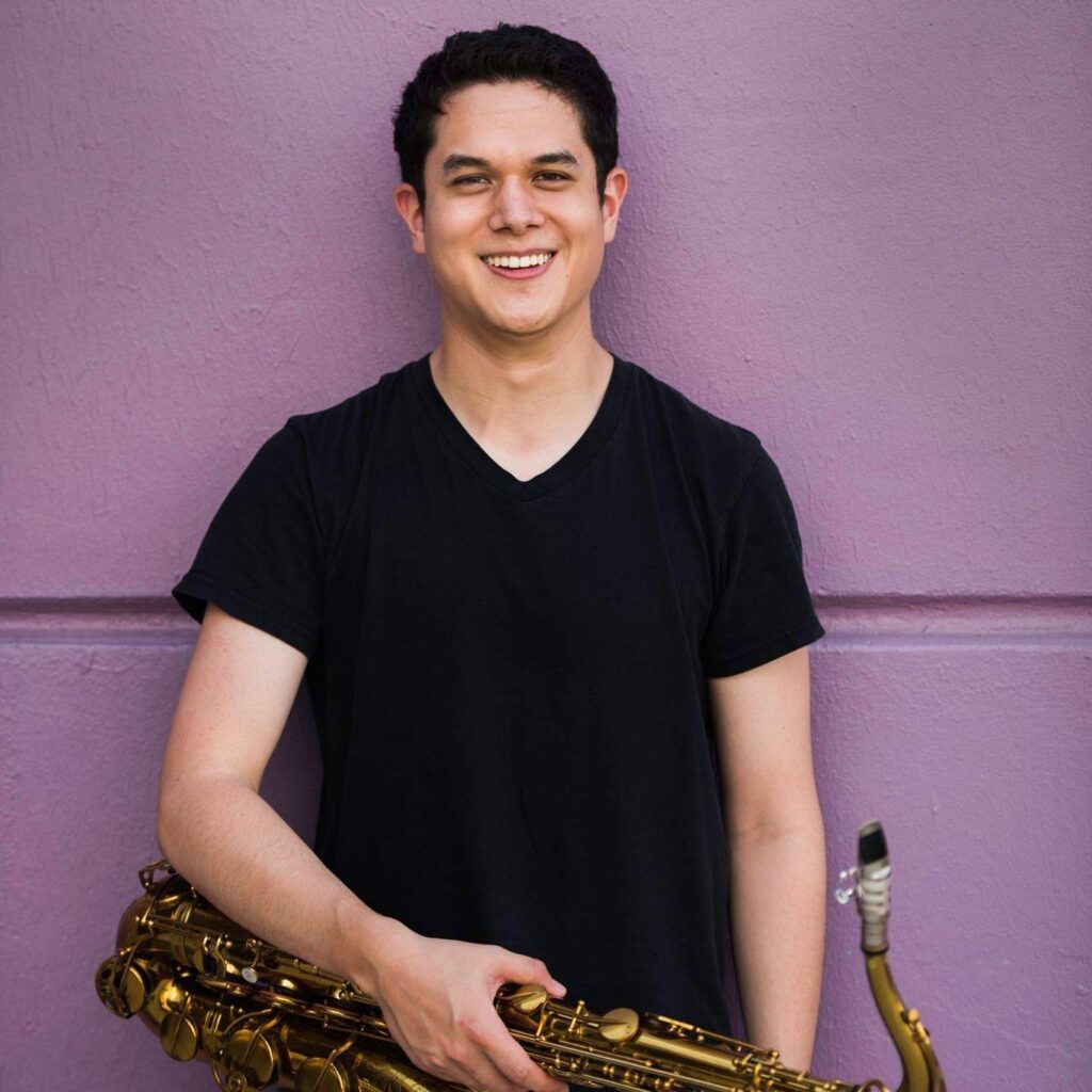 Man with dark curly hair in black v-neck shirt holding a saxophone in front of a purple wall