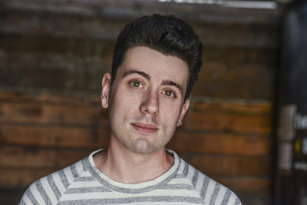 Image of man with dark quiffed hair and striped white and gray shirt in front of a brick wall