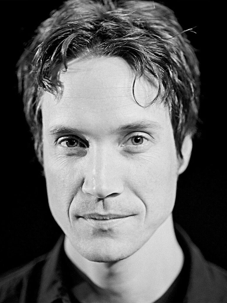 Black and white image if a man with short dark hair and black collar top