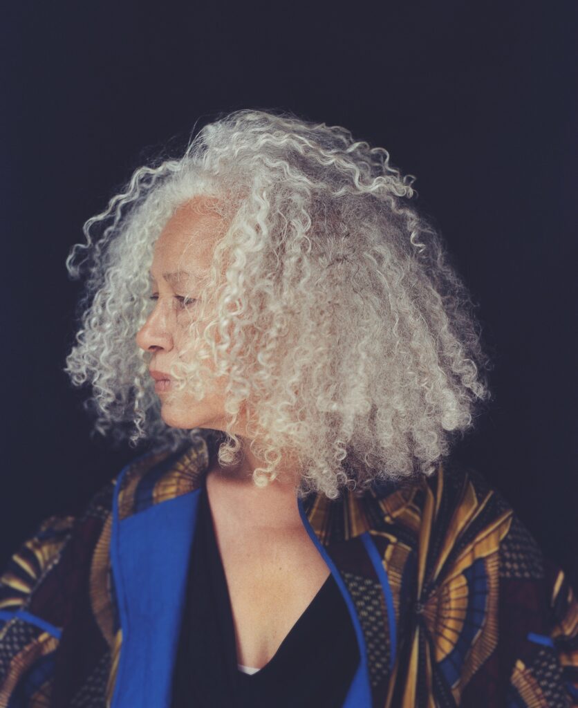 Woman with silver afro wearing a black shirt and a gold-black-blue patterned coat