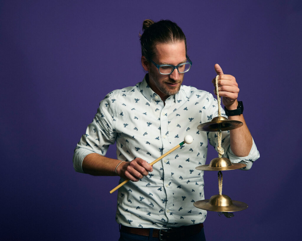 Jamie Drake holding a chain of cymbals and a mallet wearing a black patterned white shirt, eye glasses, hair in a bun in front of a purple background