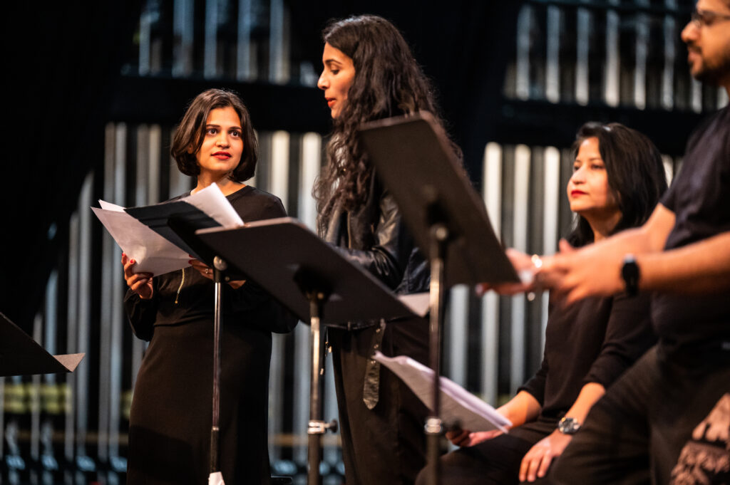 Two women with dark hair wearing all black reading off scripts on the stage.