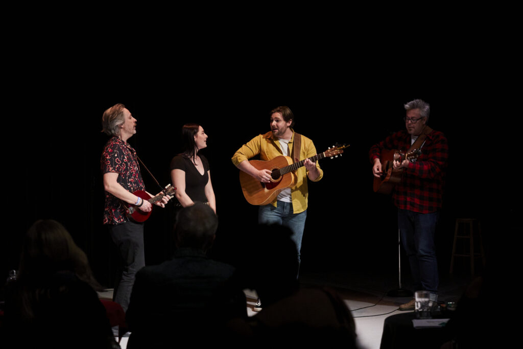 Four people in front of a dark studio theatre audience playing guitars and singing