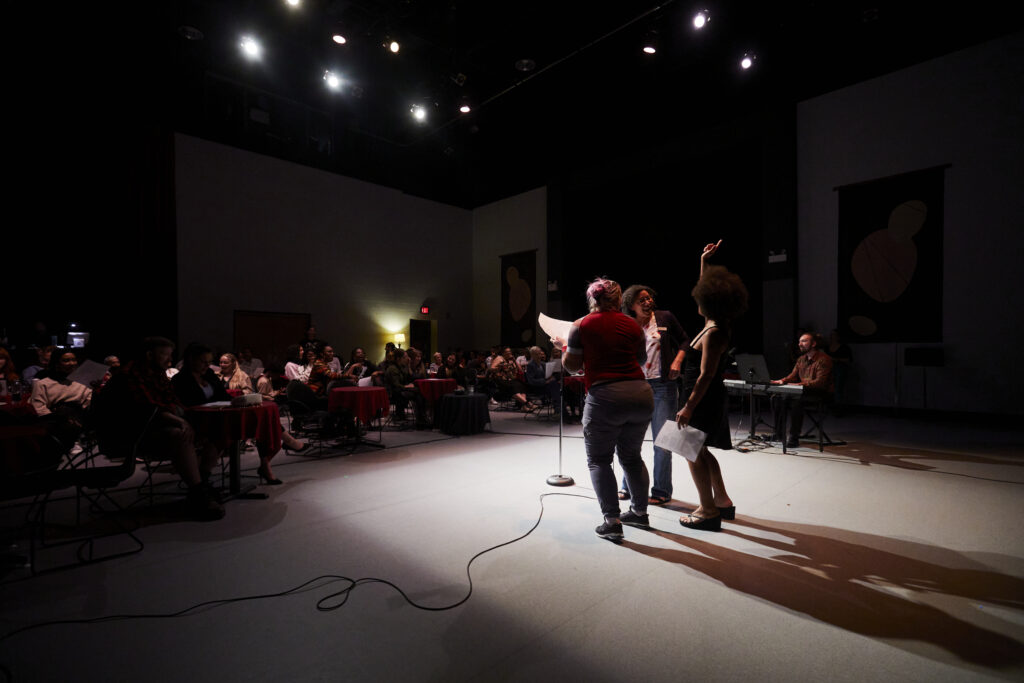 Three people laughing and reading off the script under stage lights in front of a seated audience in a dark studio theatre