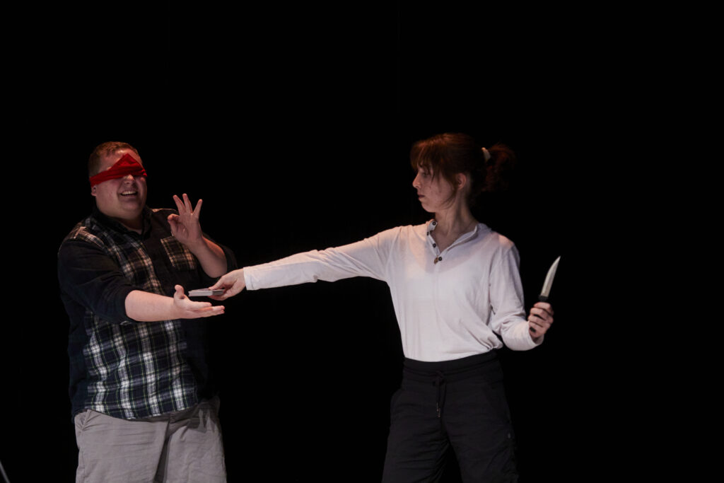 One male figure wearing a red blindfold with hand out under another female presenting figure's hand and wearing a white shirt holding a knife in the other hand.