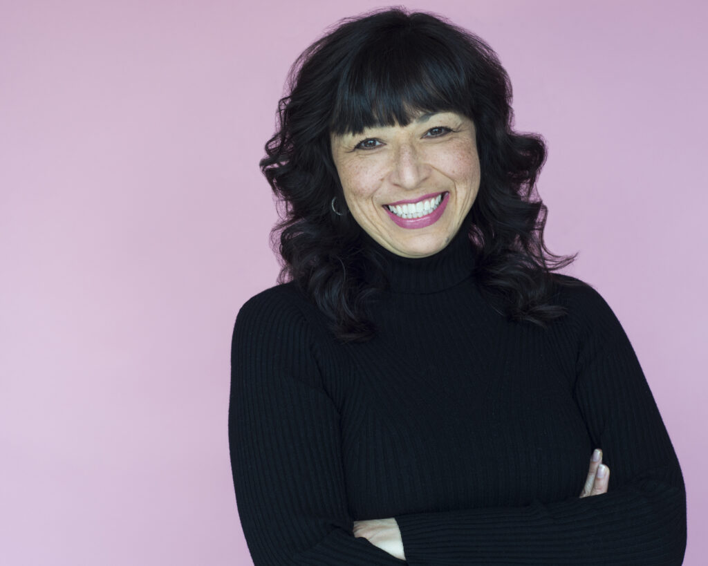 Woman with dark layered wavy hair, wide grin and wearing a black turtleneck with arms crossed in front of a pink background