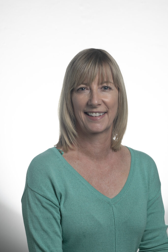 Person with blonde bangs and shoulder length straight hair wearing a blue-green v-neck top in front of a white background.