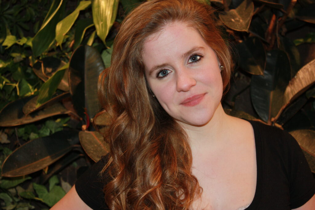 Female presenting person with long reddish-brown curled hair over right shoulder and wearing black short-sleeve top in front of green leaves.