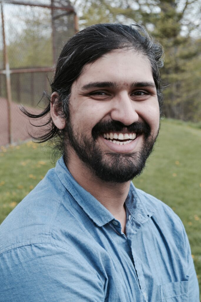 Male presenting portrait of a person with dark hair, mustache and beard wearing a blue collar shirt at a park