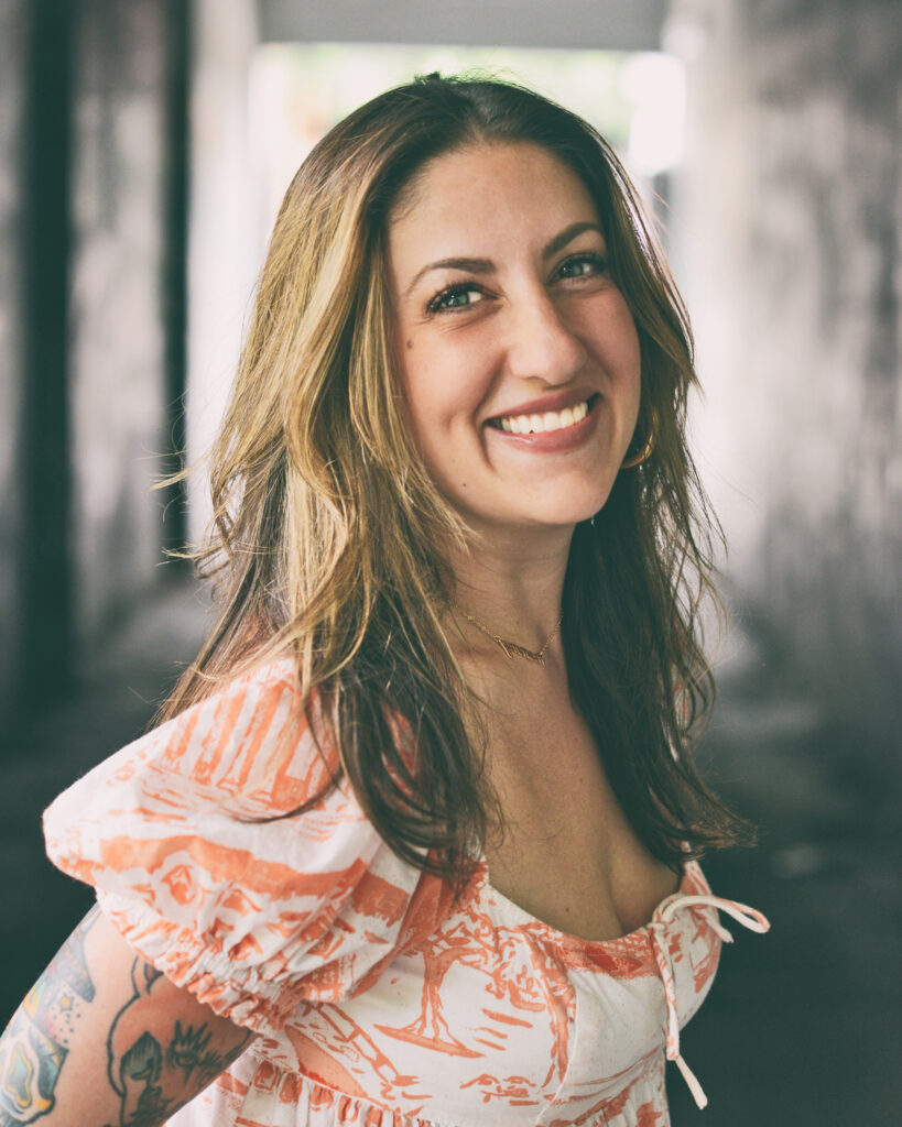 Portrait of a woman with long waved light brown hair wearing a white and peach dress standing in a tunnel.