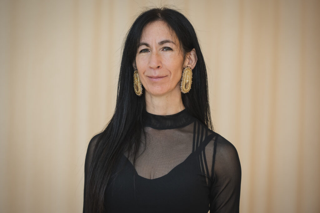 Portrait of a woman with long straight dark hair wearing a mesh black top over a black tank and large gold earrings in front of a gold background.