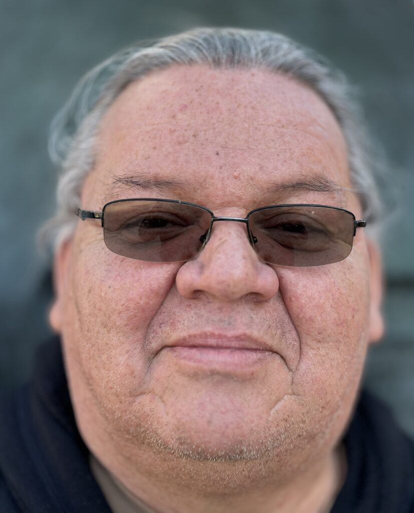 Close-up portrait of a person wearing thin rounded sunglasses, gray hair tied to the back, and a black top in front of a concrete wall.