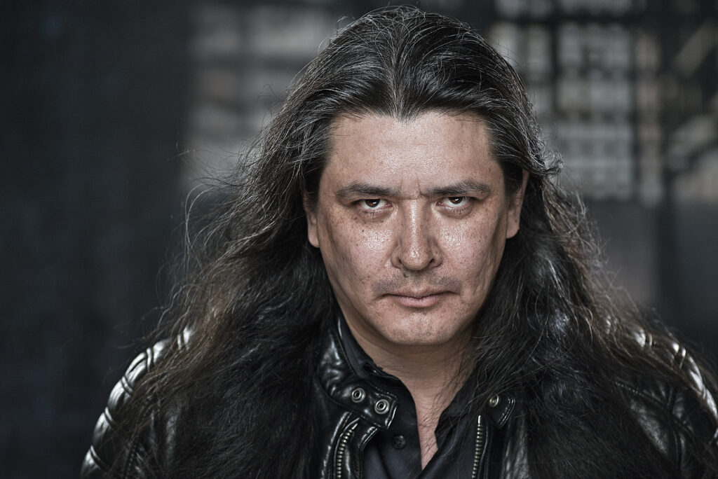 Person with long dark wavy hair, wearing a leather jacket and standing in from of a blurred dark background with window light.