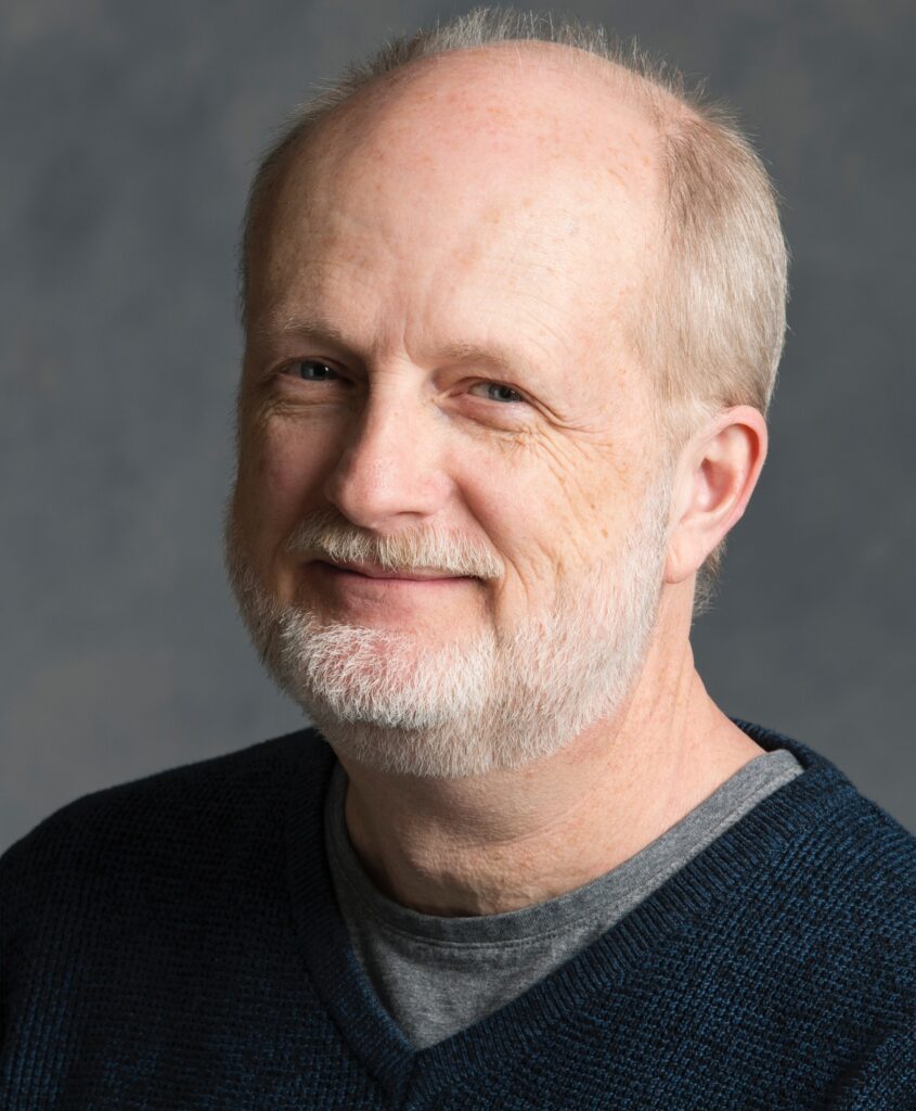 Portrait of a man with gray hair and beard wearing a black shirt over a gray shirt in front of a gray backdrop.