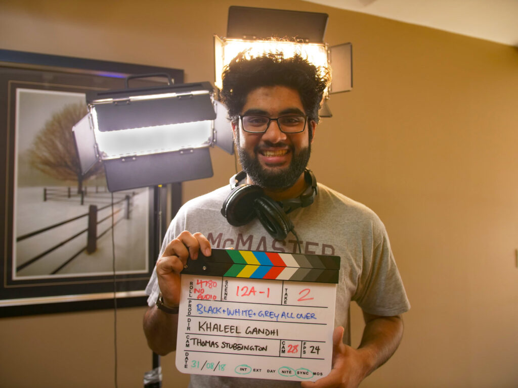 Image of a person with tall dark straight hair standing in front of studio lights, wearing a gray shirt headphones around neck and holding a clapperboard.