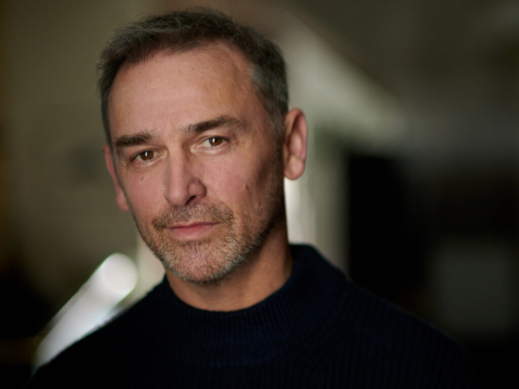 Portrait of a person with short dark gray hair wearing black turtleneck in a dim room behind him.