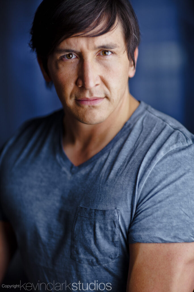 Portrait of a man wearing a blue shirt with side swept brown hair in front of a blue studio backdrop.