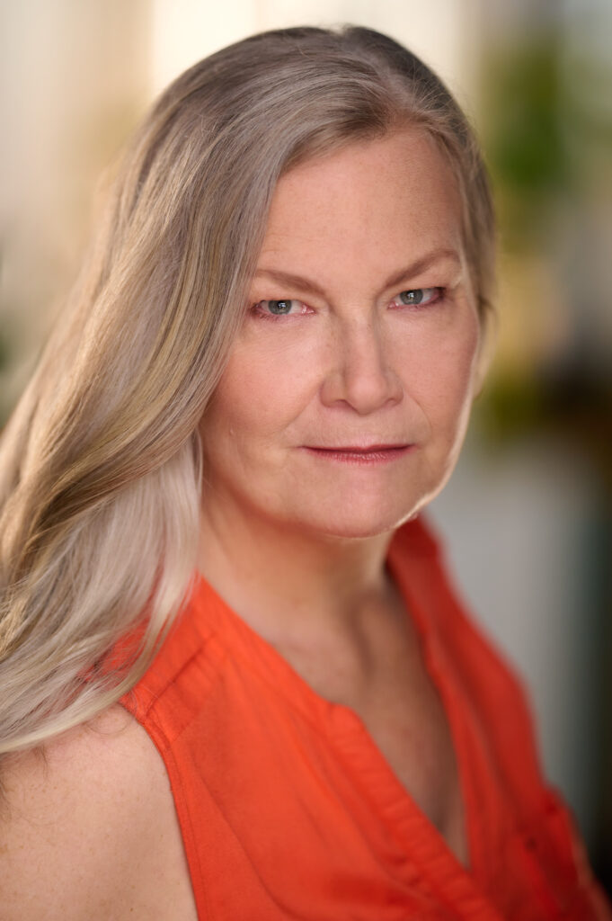 Portrait of a woman with straight long blonde hair wearing an roange sleeveless top with a blurred background behind her.