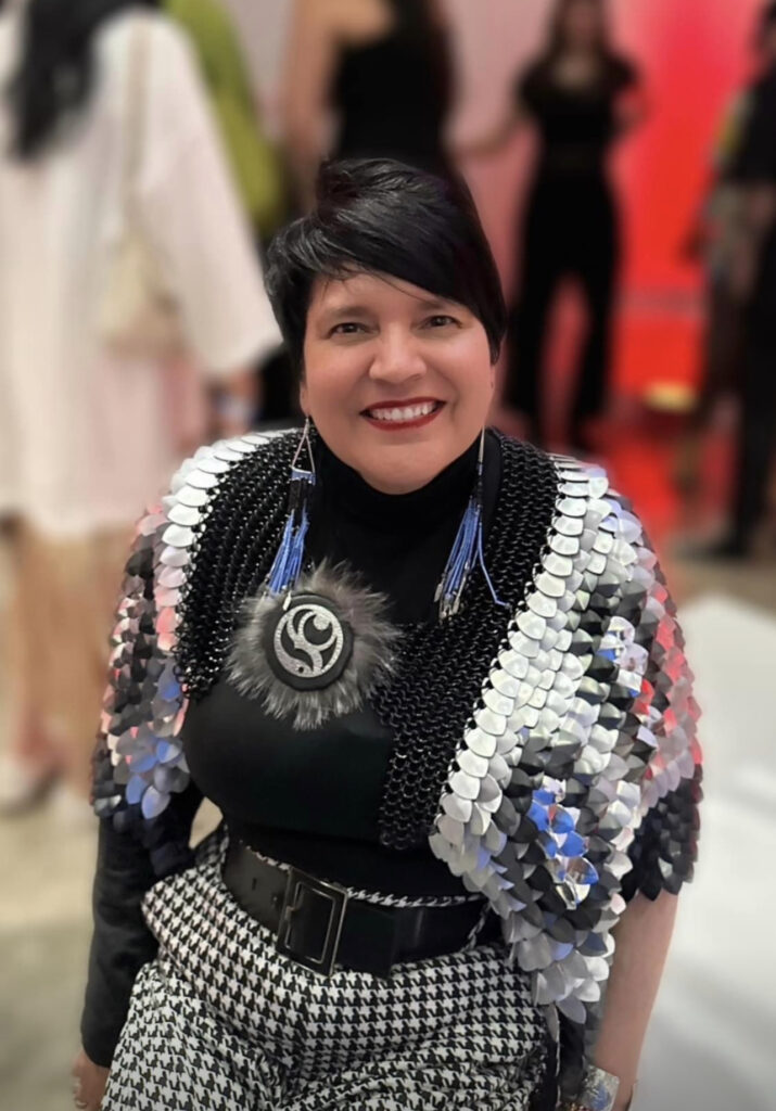 Portrait of a person with short side swept black hair wearing beaded coat and jewelry over a black top, blurred background of other people