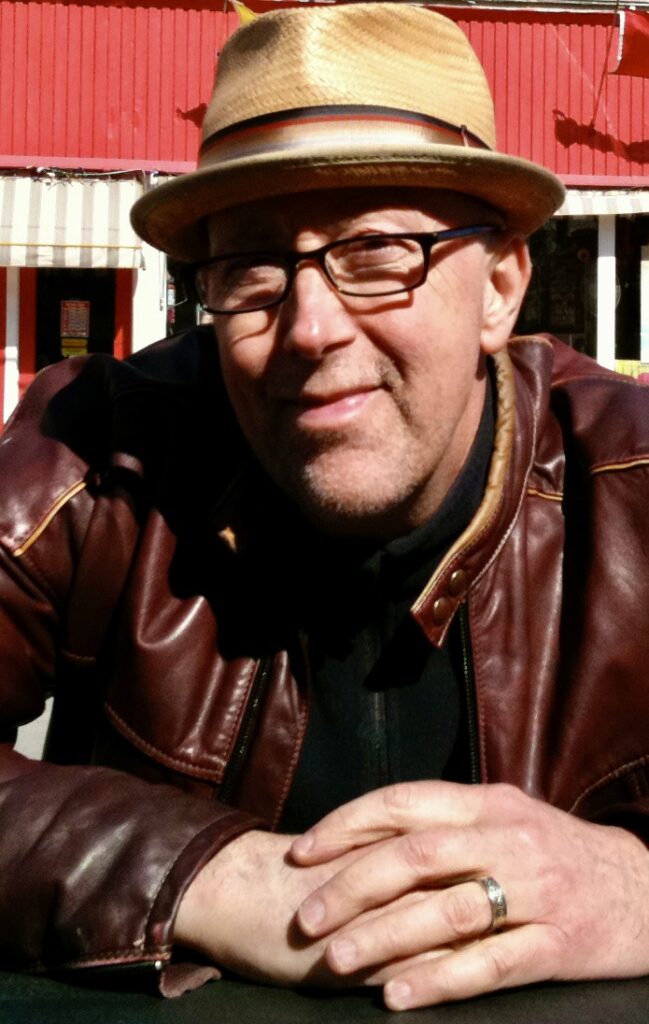 Portrait of a person with cowboy hat, brown leather jacket and eyeglasses in the sun with a red and white building in the back.