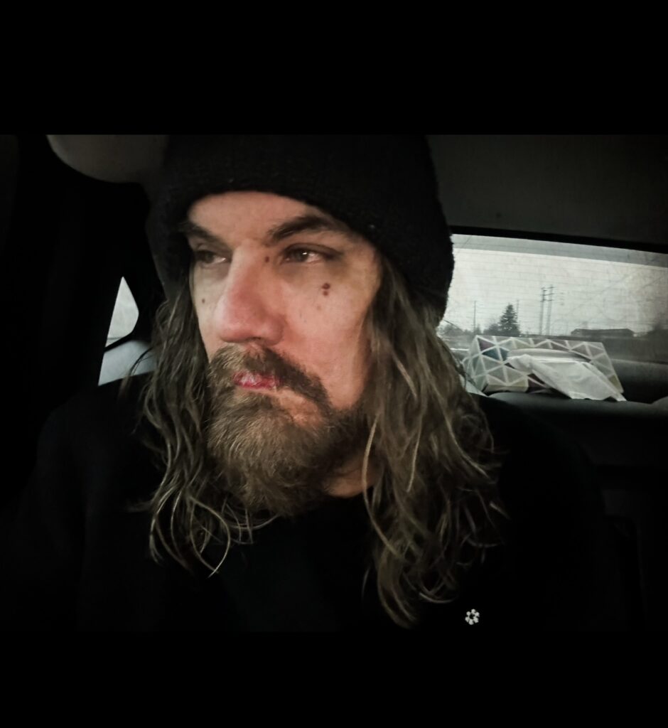 Tom in the back seat of a car wearing a black toque and black top, with long dark wavy hair and mustache and beard.