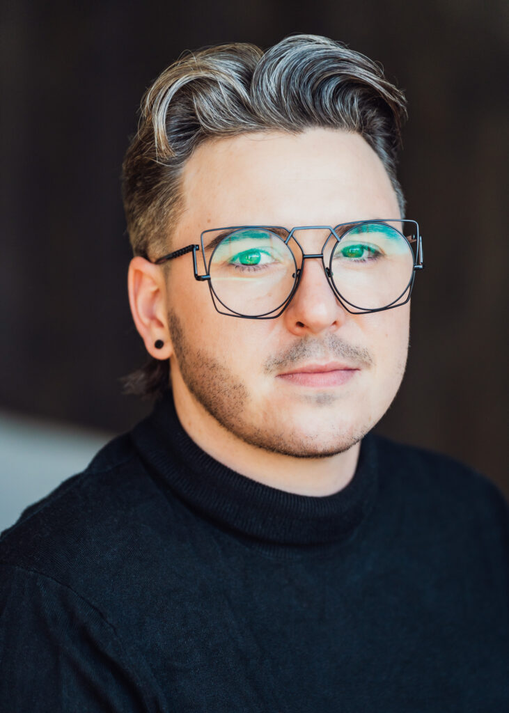 Portrait of a person wearing glasses, black stud earring, black turtleneck and quaffed and curled blond hair in front of a dark and blurry background