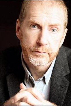 Man with thin red hair and boxed beard wearing a gray suit jacket with blue collared shirt